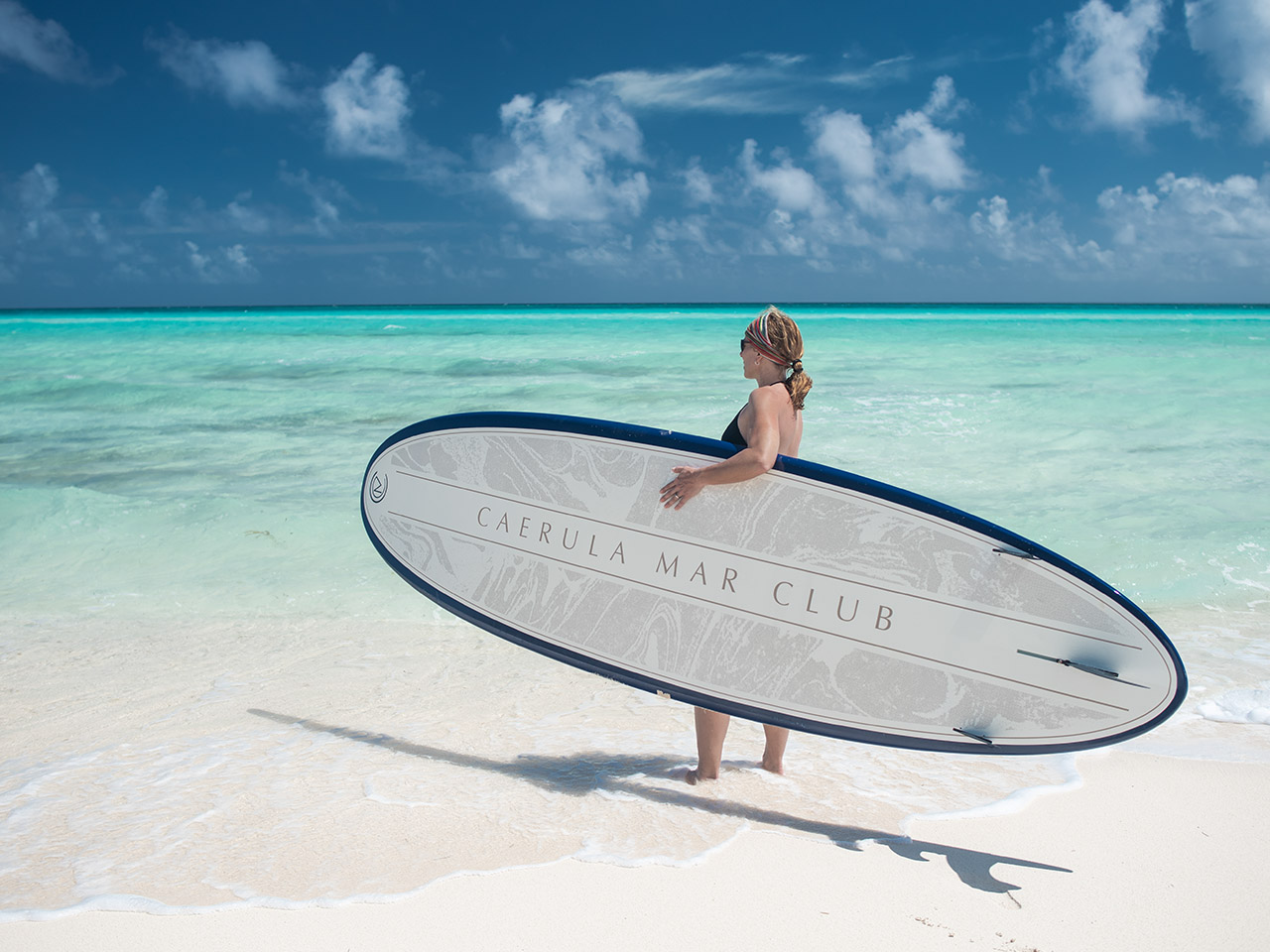 Guest with paddleboard on sandy beach at Caerula Mar Club, South Andros, Bahamas; turquoise water and blue sky in background.
