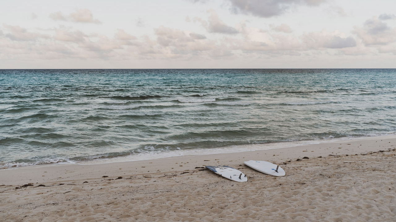 Surfboards on sandy beach by shoreline at Caerula Mar Club, luxury resort in South Andros, Bahamas; gentle waves and cloudy sky.