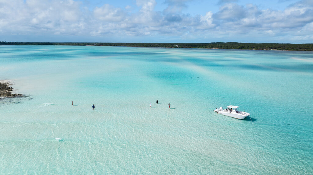 A white boat floats on turquoise water as guests wade nearby at Caerula Mar Club, with lush shoreline under a partly cloudy sky.