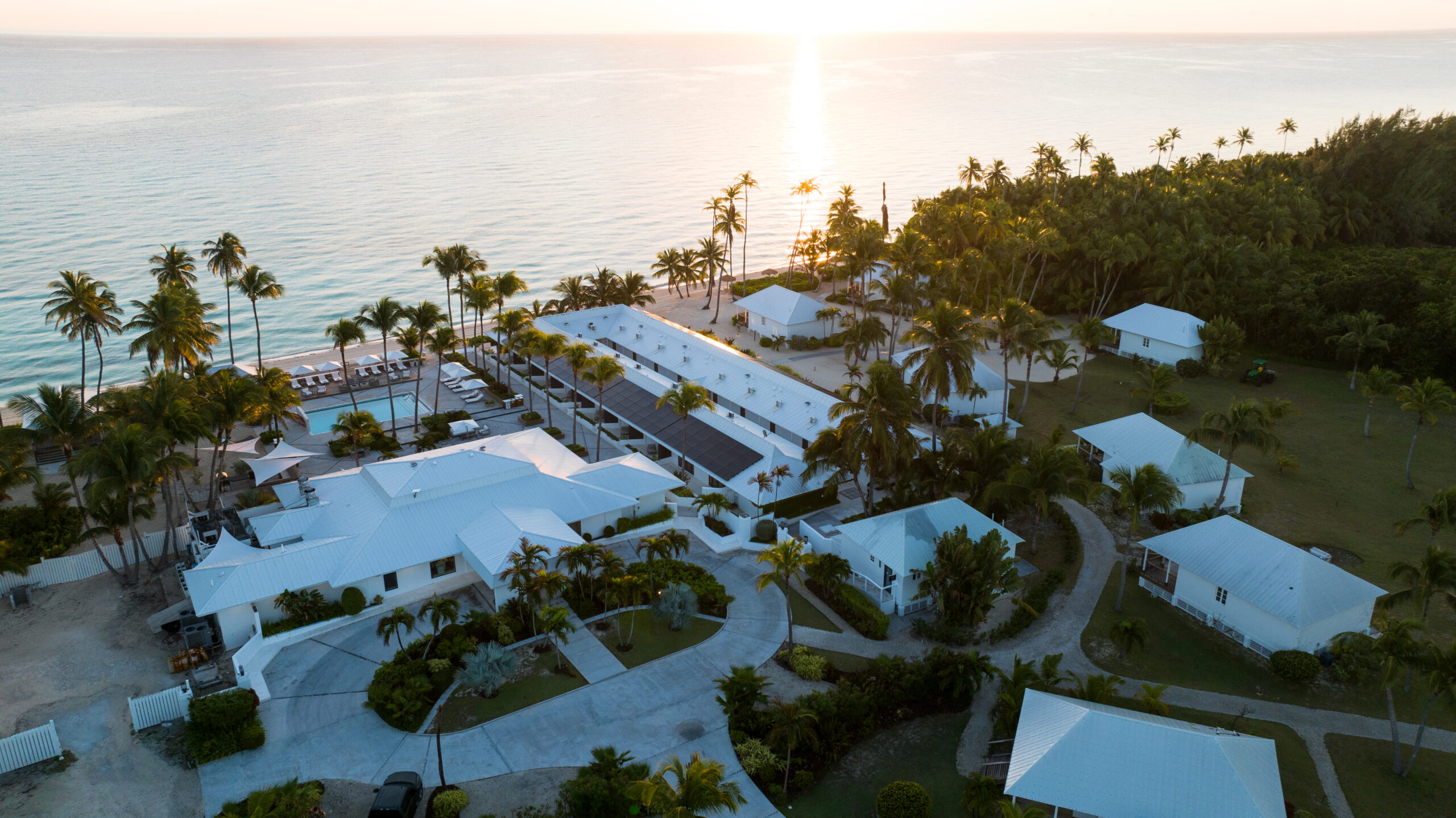 Aerial view of Caerula Mar Club on South Andros Island in The Bahamas, featuring elegant white buildings, swaying palm trees, and a pristine sandy beach at sunset beside tranquil ocean waters.