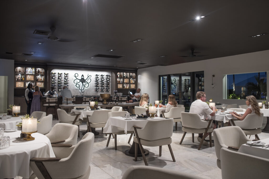 Elegant restaurant interior at Caerula Mar Club, a luxury boutique resort on South Andros Island in The Bahamas, featuring white tables with cushioned chairs, guests dining, attentive waitstaff serving, wine bottles displayed on the wall, modern decor with dim lighting, and large windows revealing dusk views of the outdoors.